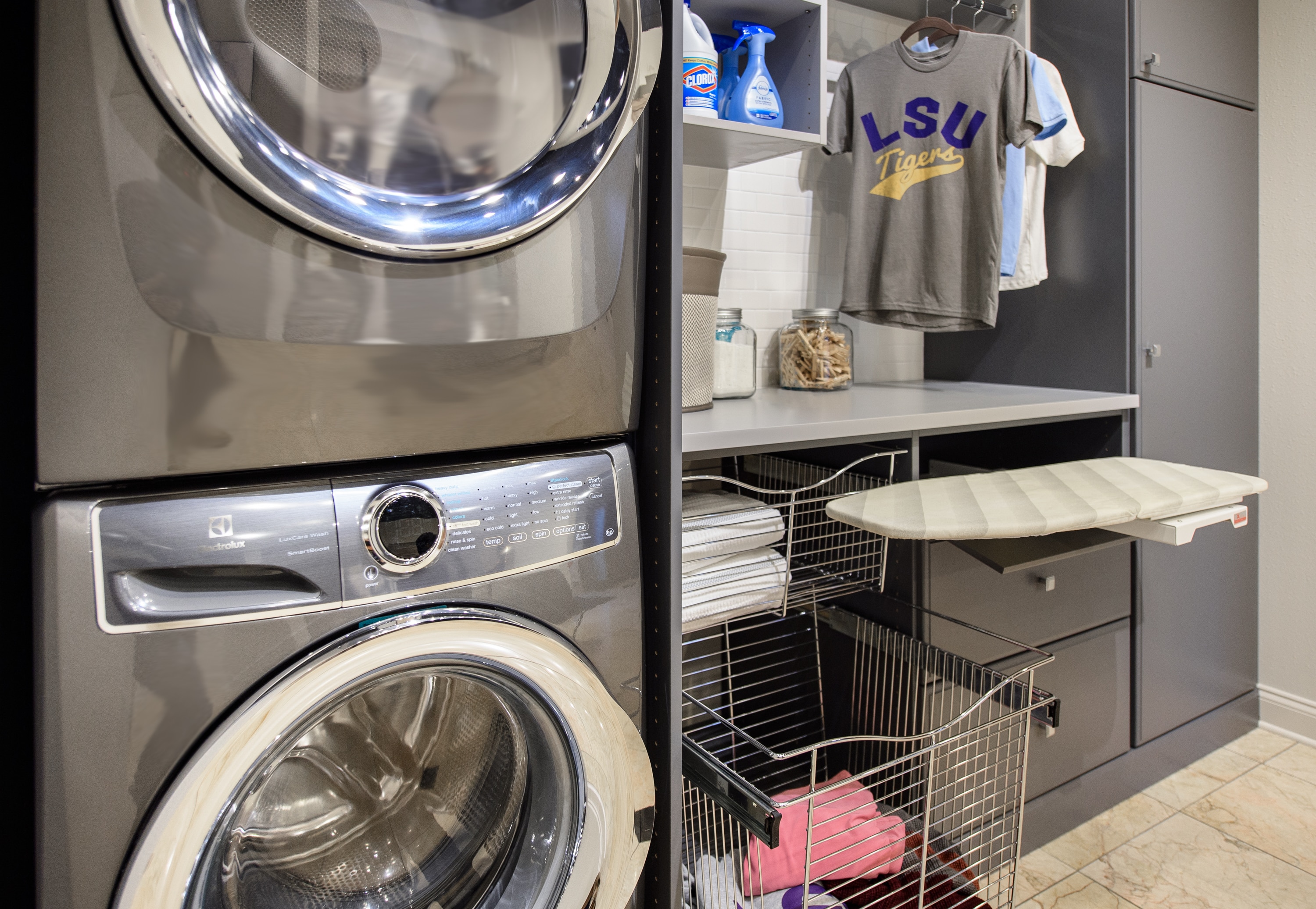 Laundry closet showing washer and dryer as well as clothing baskets, ironing board and shirts on rack